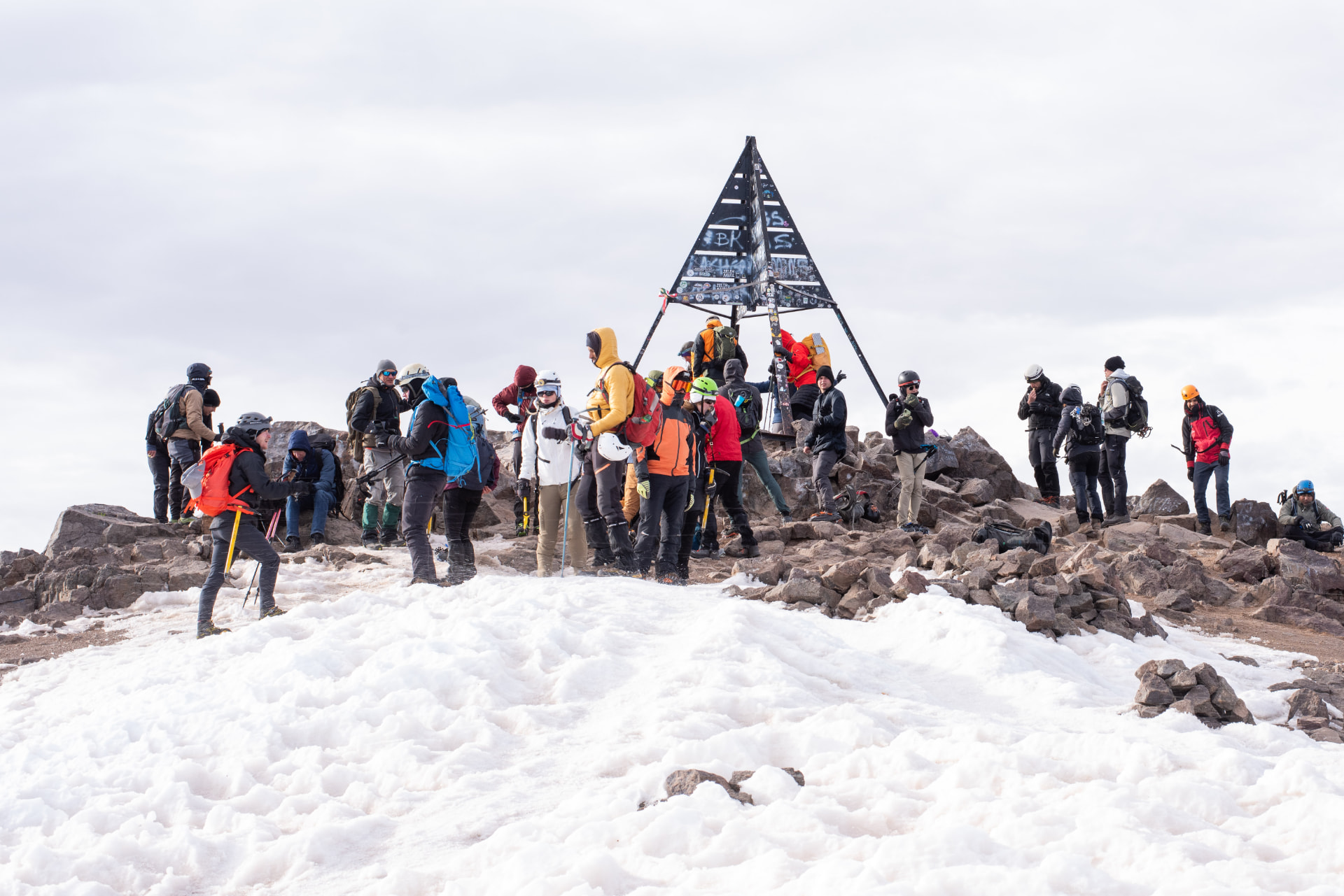 Group of hikers gathered around the summit marker on Mount Toubkal with snow and rocky peaks