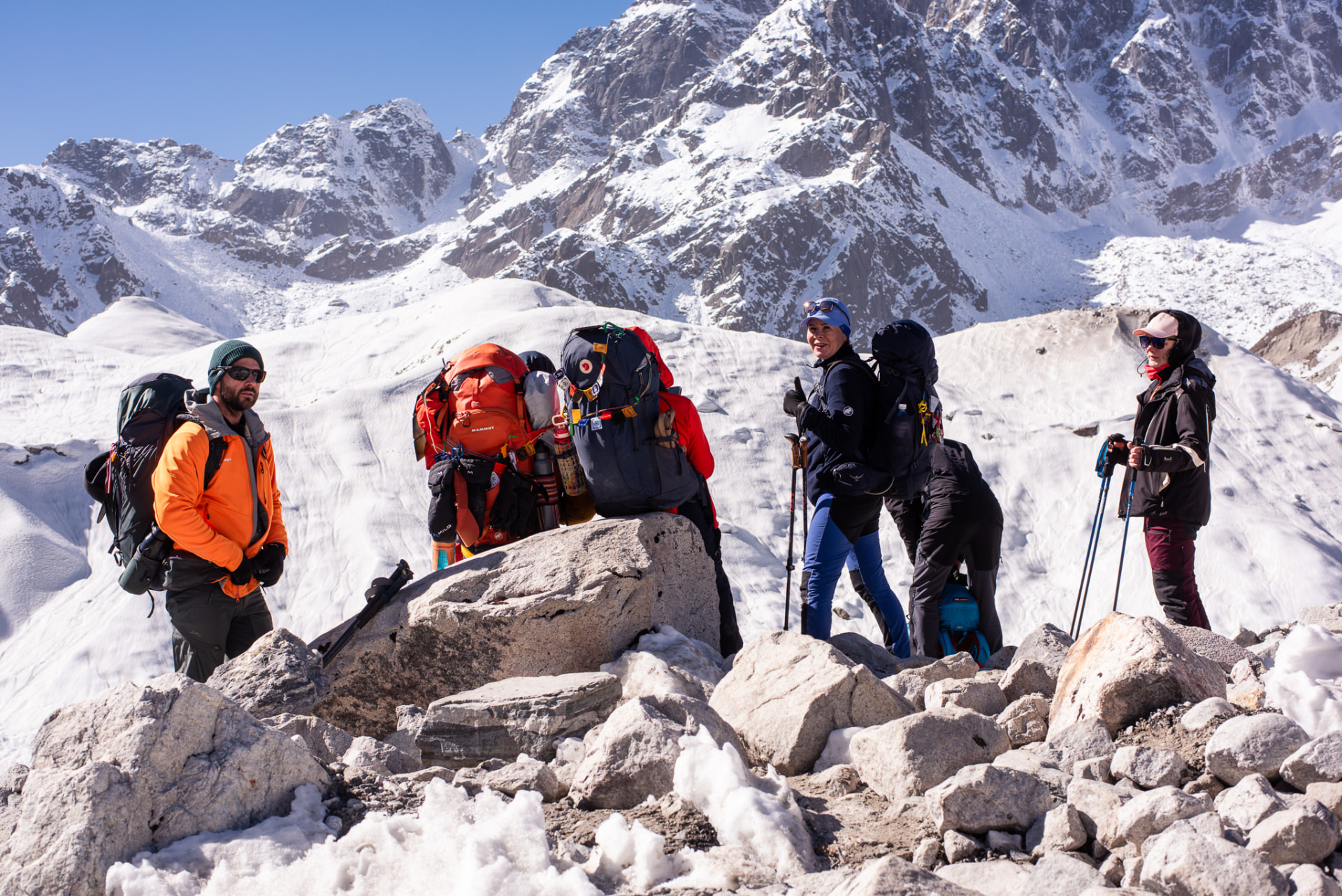 Group of hikers with large backpacks resting on rocky Himalayan terrain with snow-covered peaks behind them